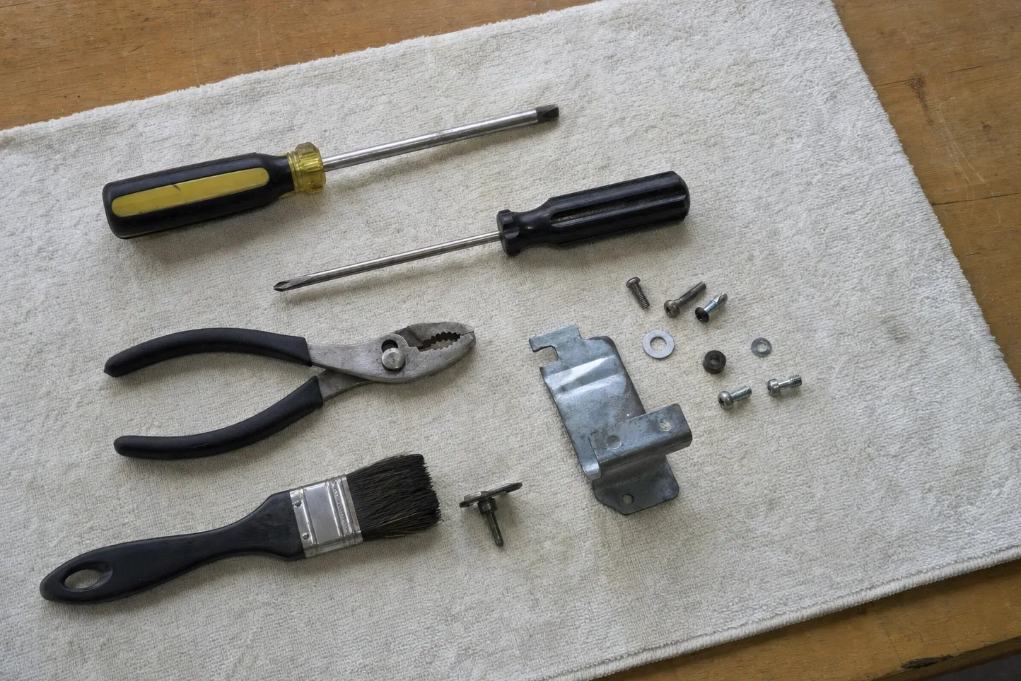Screwdrivers, slip-joint pliers and a bristle brush on a towel beside a galvanised bracket, washers and a push rivet